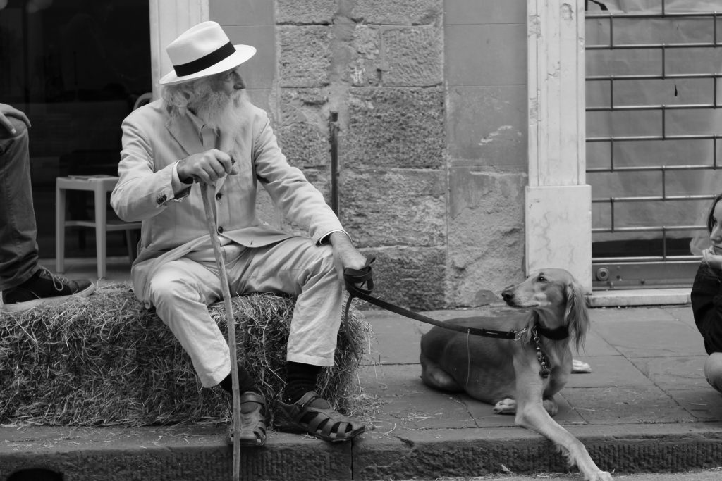 Vanha parrakas mies istuu heinäpaalin päällä ja pitää koiraa hihnan päässä.